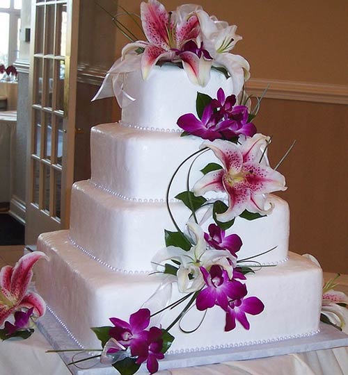 Square four tier white fondant wedding cake decorated with tropical flowers, like purple orchids, white oriental lillies and pink tiger lillies